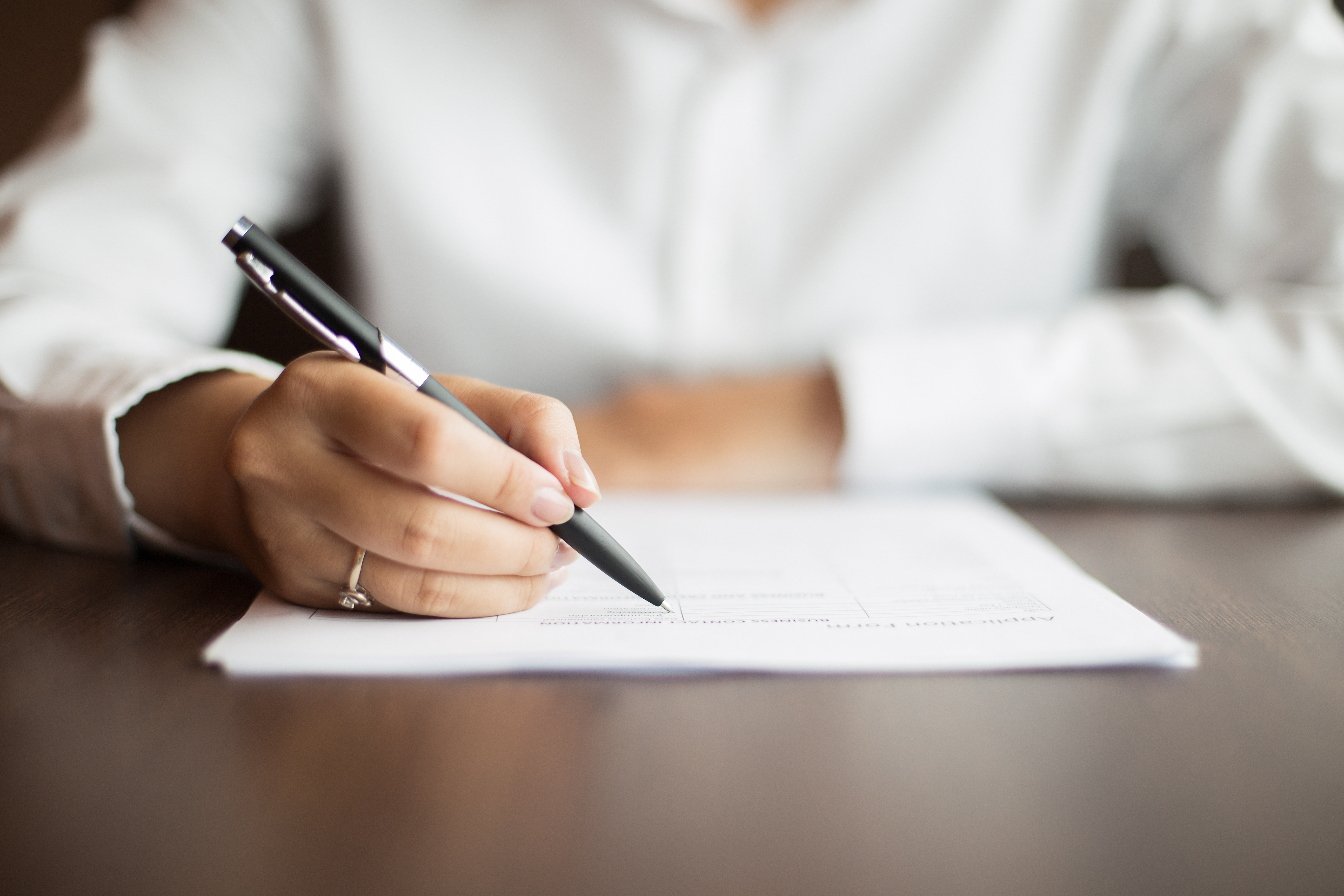 Hand of young businesswoman wearing ring and sitting at table writing on document with pen in office